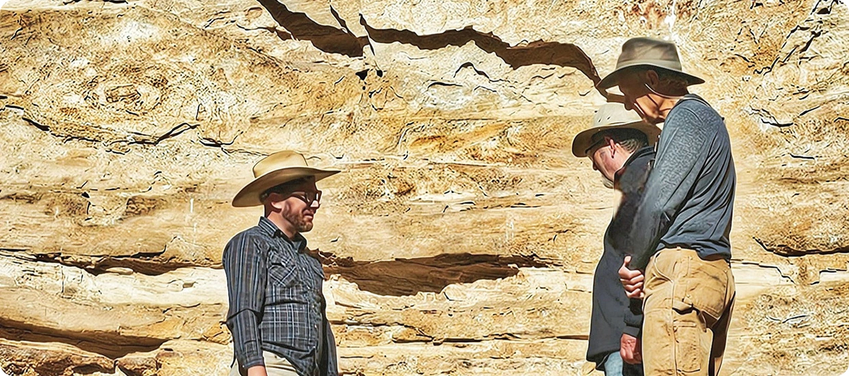 Photograph of three people standing in front of a tan sandstone wall.